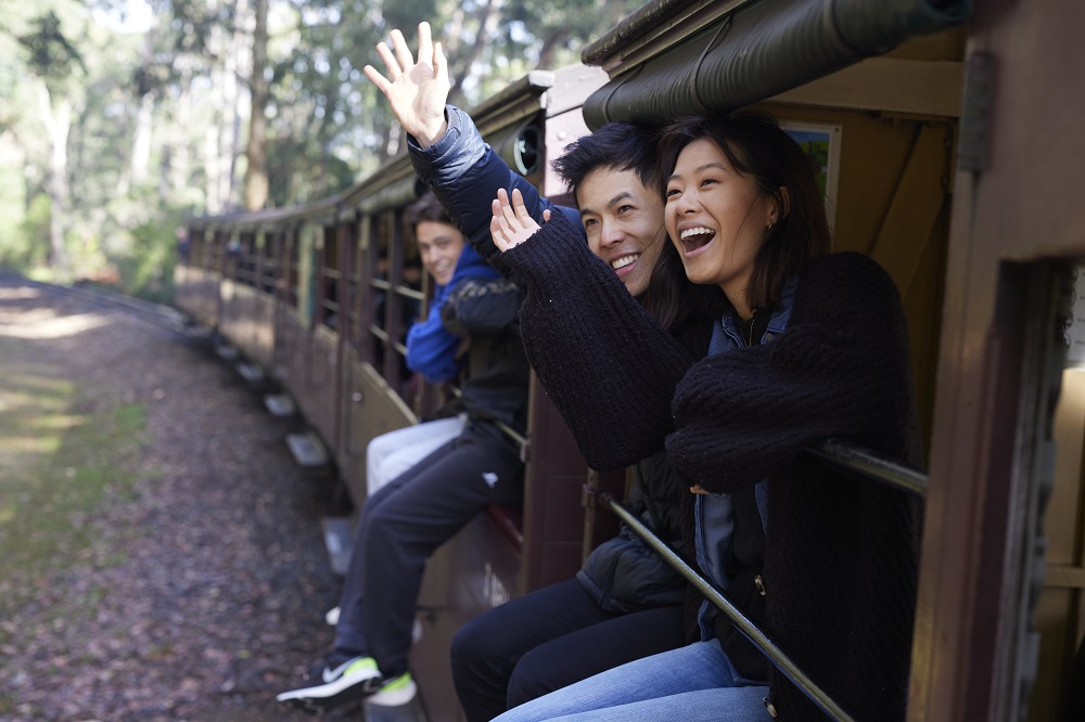 A young couple are sitting on the carriage sills of an open side carriage. They are waving and smiling to someone off-camera.